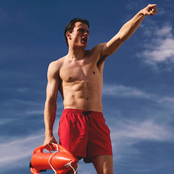 HUNQ - #004 Lifeguard zeigt muskulösen Mann in roter Badehose mit Rettungsboje vor bewölktem Himmel