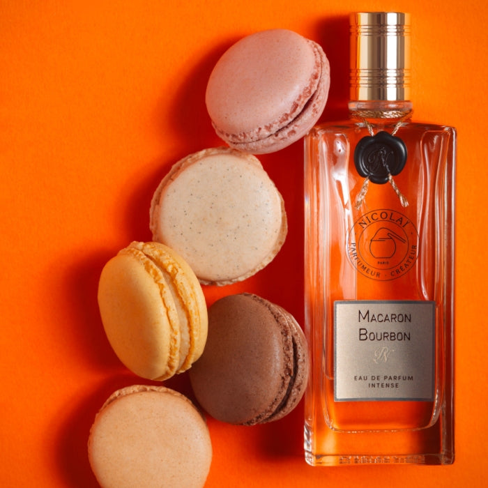 Nicolaï - Macaron Bourbon Intense Parfümflakon neben fünf bunten Macarons auf leuchtend orangefarbenem Hintergrund
