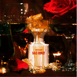 BORNTOSTANDOUT - Mad Honey Parfümflakon mit tropfendem Honig, umgeben von roten Rosen und Glaswein im warmen Licht