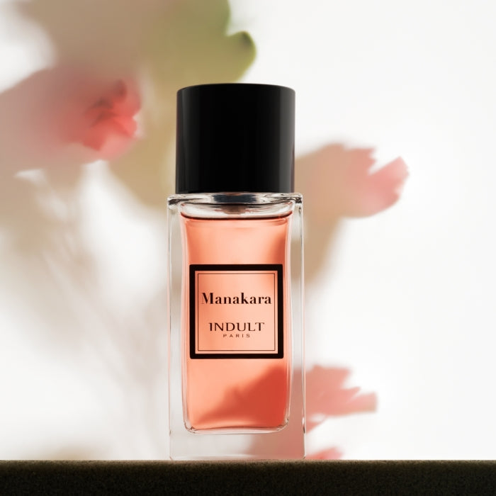 Indult - Manakara Parfumflakon mit rosafarbenem Duft und schwarzem Verschluss vor zart verschwommenen Blüten im Hintergrund
