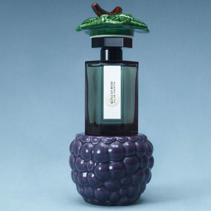 L'Artisan Parfumeur - La ollection - Mûre et Musc Parfumflakon mit kunstvoll geformtem Beerenverschluss vor blauem Hintergrund