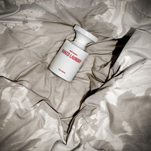 BORNTOSTANDOUT - Naked Laundry Parfumflakon in weiß mit roter Schrift auf zerknittertem, feuchtem Stoff in sinnlicher Atmosphäre