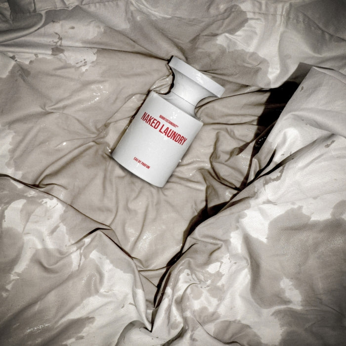 BORNTOSTANDOUT - Naked Laundry Parfumflakon in weiß mit roter Schrift auf zerknittertem, feuchtem Stoff in sinnlicher Atmosphäre