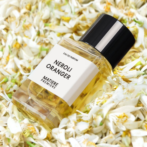 Matière Première - Neroli Oranger zeigt klaren Flakon mit hellem Parfum vor Blüten der Orangenblüte