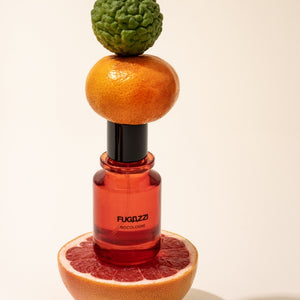 Fugazzi - Nocologne roter Flakon balanciert zwischen Grapefruithälfte, Mandarine und grünem Bergamottenfruchtkörper