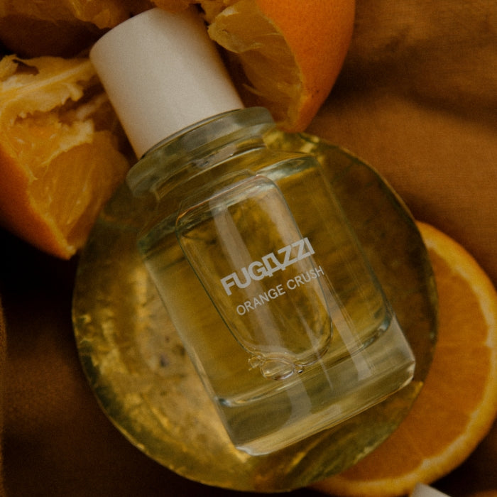 Fugazzi - Orange Crush Parfumflakon mit rundem Design und elfenbeinfarbenem Verschluss vor Orangenspalten auf dunklem Hintergrund