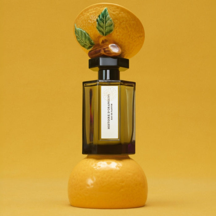 L'Artisan Parfumeur - La Collection - Histoire d'Orangers Parfumflakon mit kunstvollem Orangen-Deckel vor gelbem Hintergrund