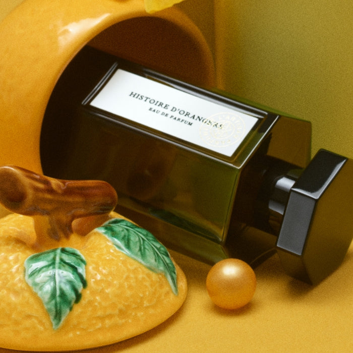 L'Artisan Parfumeur - La Collection - Histoire d'Orangers Parfumflakon in dunklem Glas neben Keramikorange vor gelbem Hintergrund