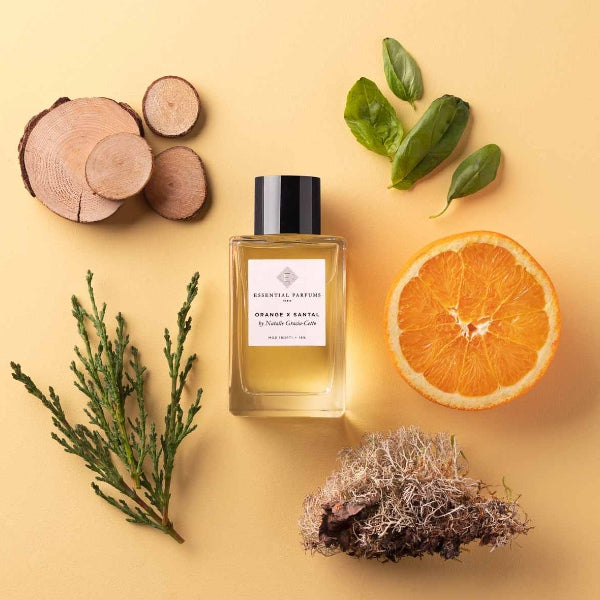 Essential Parfums - Orange X Santal Parfumflakon mit Holzscheiben, Orangenhälfte und grünen Blättern auf beigem Hintergrund