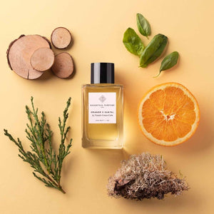Essential Parfums - Orange X Santal Parfumflakon mit Holzscheiben, Orangenhälfte und grünen Blättern auf beigem Hintergrund