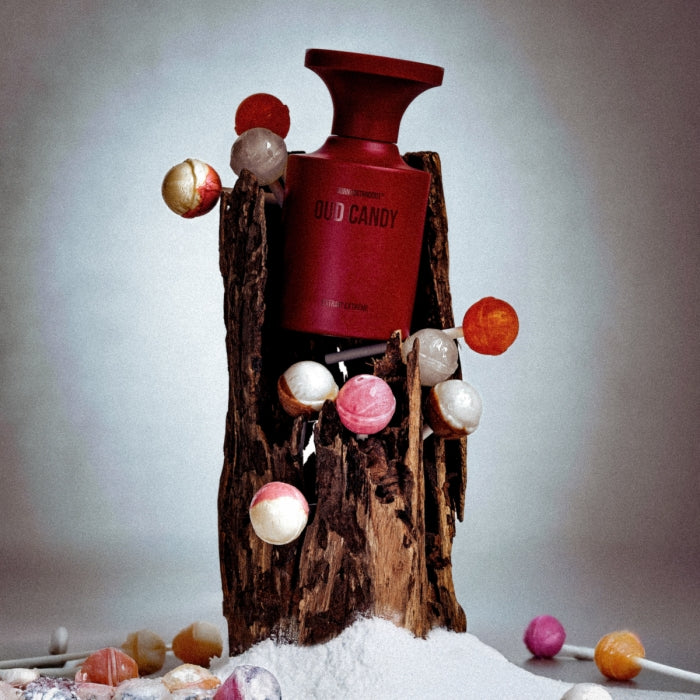 BORNTOSTANDOUT - Oud Candy Flakon in mattem Dunkelrot, inszeniert mit Holzstück und bunten Lutschern auf weißem Untergrund
