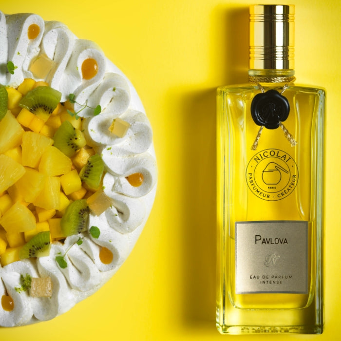 Nicolaï - Pavlova Intense Parfümflakon neben exotischem Pavlova-Dessert auf lebendig gelbem Hintergrund