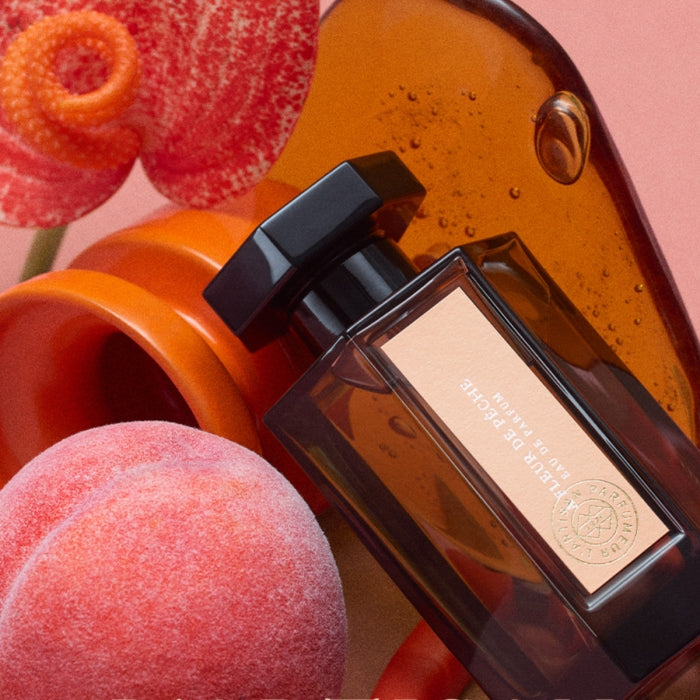 L'Artisan Parfumeur - La Collection - À Fleur de Pêche zeigt Flakon vor Pfirsich und orangefarbenen dekorativen Elementen