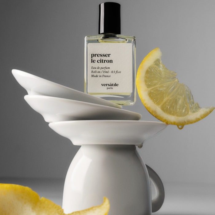 Versatile - Presser le Citron Roll-on Parfumflakon balanciert auf weißem Geschirr mit frischer Zitronenscheibe nahe