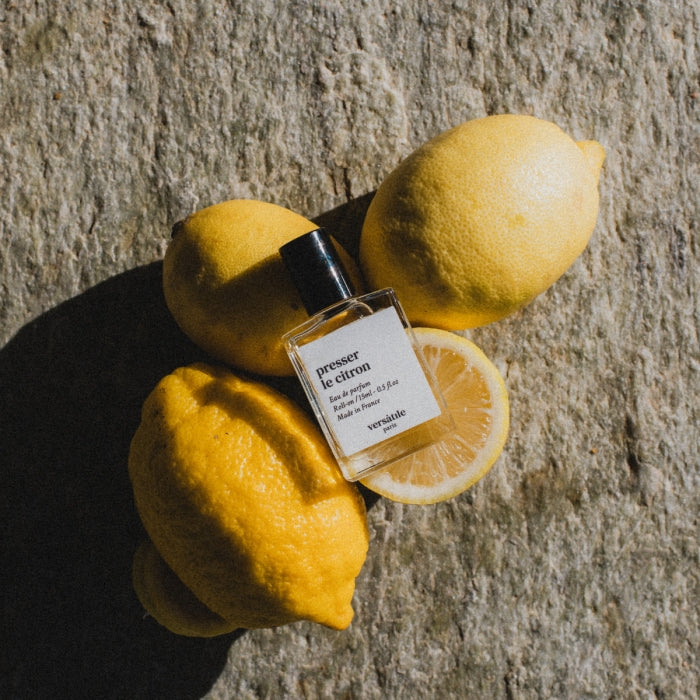 Versatile - Presser le Citron Parfumflakon zwischen frischen Zitronen auf rustikalem steinernen Untergrund in natürlichem Licht
