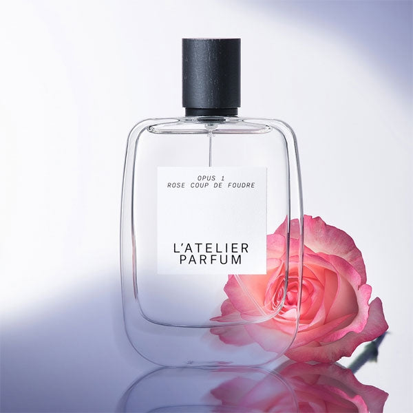 L'Atelier Parfum - Opus 1 - Rose Coup de Foudre klare Flasche mit schwarzem Verschluss neben zarter rosa Rose auf hellem Hintergrund