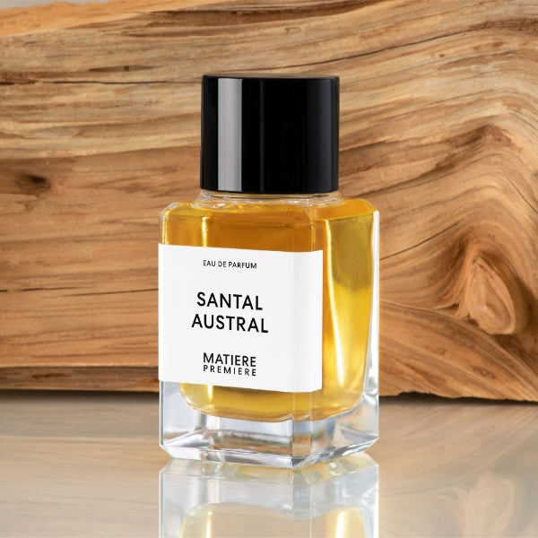 Matière Première - Santal Austral zeigt klaren Flakon mit schwarzem Deckel und goldgelbem Duft vor Holzstruktur