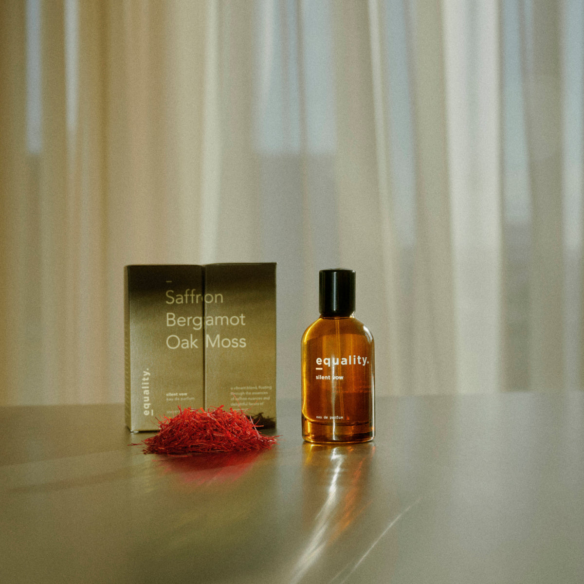 equality. fragrances - silent vow Parfumflakon in bernsteinfarbenem Glas mit Verpackung und Safran vor transparentem Vorhanglicht