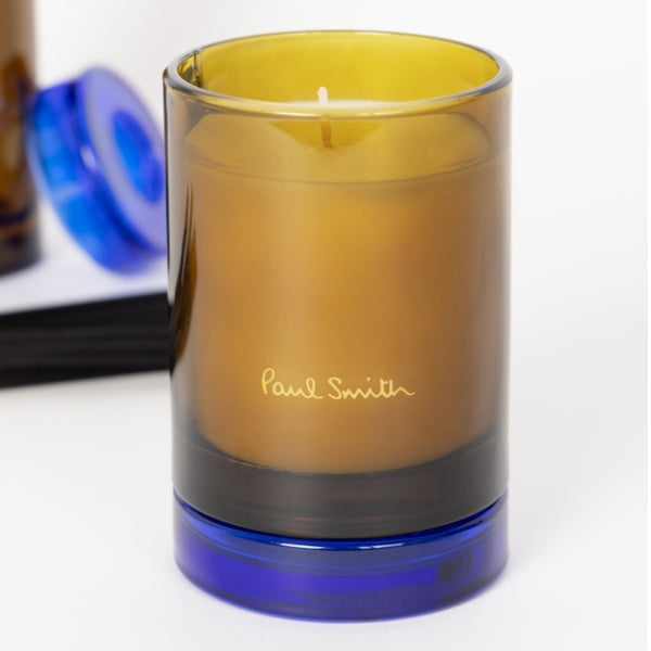 Paul Smith - Storyteller - Duftkerze im bernsteinfarbenen Glas mit blauem Sockel und dezenter Signatur auf hellem Hintergrund