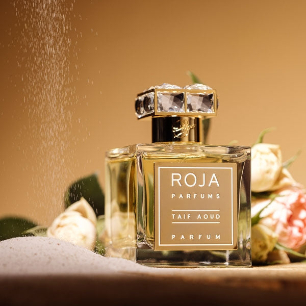 Roja Parfums - Taif Aoud Parfum Flakon mit goldfarbenem Etikett und luxuriösen Schmuckdetails vor Blumen und Sand