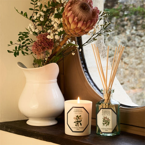 Carrière Frères - Tiare - Gardenia Tahitensis - Duftkerze im weißen Glas mit elegantem Etikett neben Blumen und Diffusor