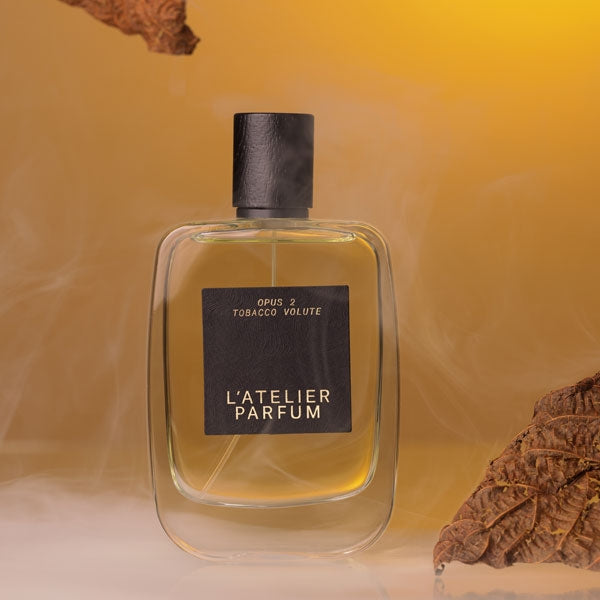 L'Atelier Parfum - Opus 2 - Tobacco Volute klarer Flakon mit schwarzem Etikett vor warmem, nebligem Hintergrund und getrockneten Tabakblättern
