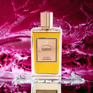 Pompeii - Valeria Parfumflakon mit goldfarbenem Verschluss und bernsteinfarbenem Duft vor pink-rotem Aquarellhintergrund