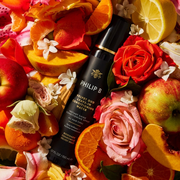 Philip B - Velvet Oud Leave-In Conditioning Water in schwarzer Sprühflasche umgeben von frischen Blüten und Früchten für seidig glänzendes Haar