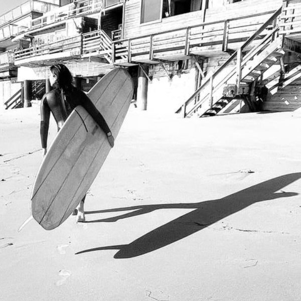 Room 1015 - Wavechild zeigt eine Surfersilhouette am Strand mit Surfbrett und langem Schatten in Schwarz-Weiß.