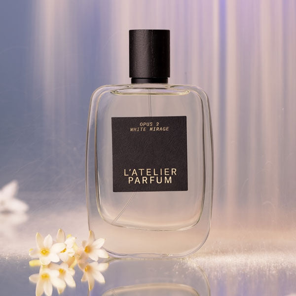 L'Atelier Parfum - Opus 2 - White Mirage klarer Flakon mit schwarzem Verschluss und weißen Blüten vor zartem Hintergrund